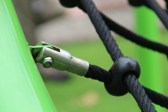 Detailed close-up of a metallic clevis joint connecting a black rope net to a green post at an outdoor playground. Concept of public facility safety standards, engineering, and maintenance.