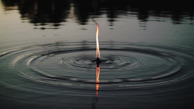 Close-up of a lit matchstick dropped into calm water creating ripples at dusk