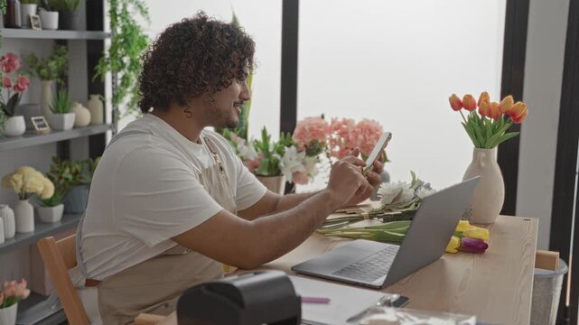Man tapping smartphone and holding stems while arranging tulips at a shop table with laptop, vase and label printer in a studio; calm focus.