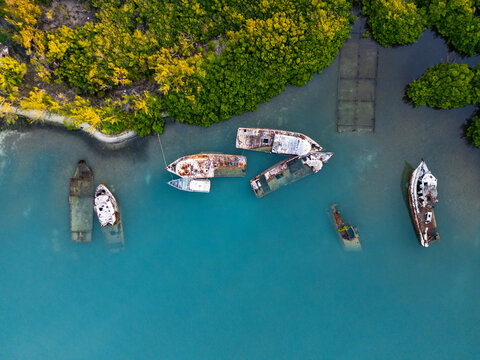 Aerial view of several rusty shipwrecks submerged in turquoise water along a lush mangrove shoreline Guanaja, Bay Islands Department, Honduras.