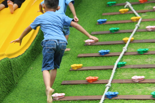 A child is climbing up a wooden obstacle course with colorful grips and a safety rope on green artificial grass. Concept of childhood physical activity, outdoor fitness, and gross motor skills develop