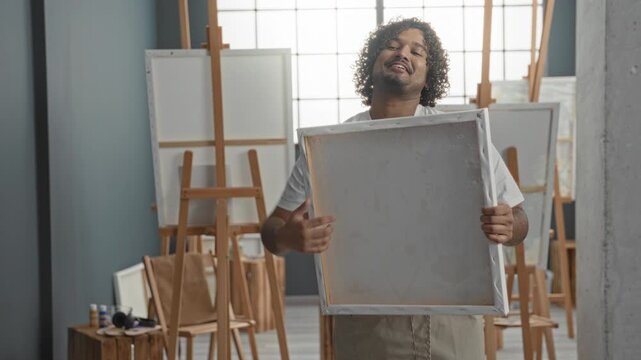 Man painter holds canvas with both hands, presenting colorful abstract painting on canvas in studio; quiet pride.