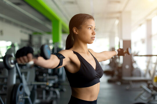 Focused young Black woman doing lateral dumbbell raises in gym, arms extended to sides, wearing black sports bra and fitness tracker armband, serious side profile, bright gym equipment background
