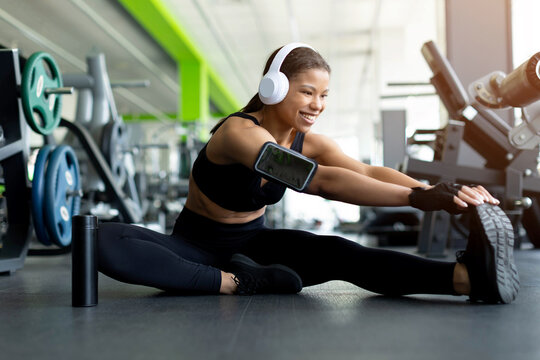 Happy young Black woman doing seated side split stretch on gym floor, smiling, wearing white headphones, black sports bra, phone armband and leggings, water bottle nearby, gym equipment background