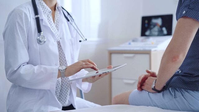 Doctor woman is taking notes on a tablet computer while a male patient describes his symptoms in clinic procedure cabinet. Medicine service concept