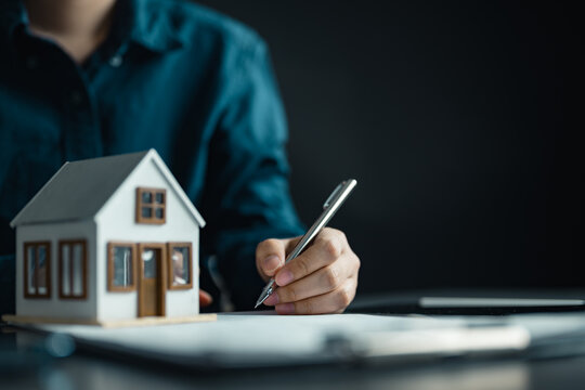 Person signing real estate contract near house model, representing home purchase, property investment, and mortgage agreement in financial planning and ownership transaction process.