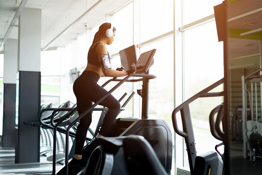 Young Black woman using stair climber machine in gym viewed from side, wearing white headphones, black sports bra and phone armband, focused expression, bright floor-to-ceiling windows in background