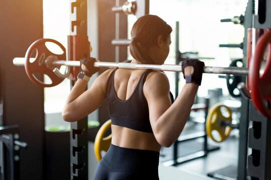 Young Black woman holding barbell across shoulders in gym squat rack, wearing black sports bra, fingerless gloves and smartwatch, preparing for barbell squat, gym equipment background, rear view