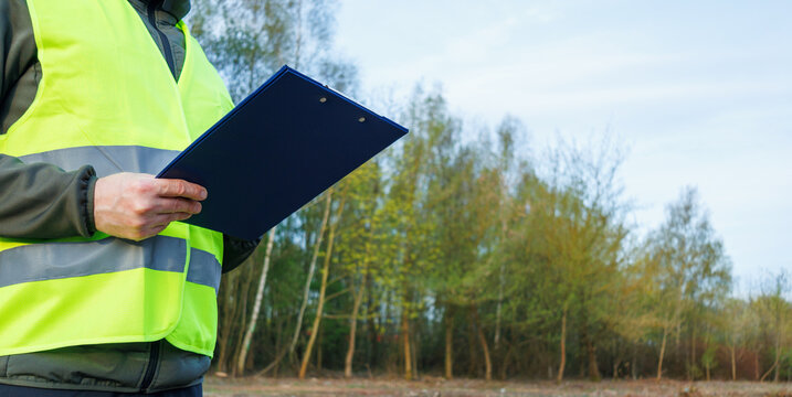 Close-up view of hands inspector holding a clipboard against a woodland background in early spring. Ecologist or site supervisor documenting environmental data during a forest inspection. 