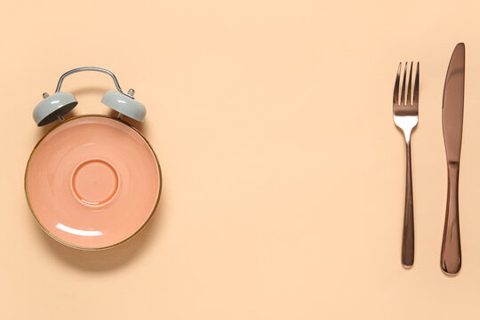 Empty plate in shape of alarm clock with cutlery on beige background. Intermittent fasting concept