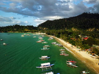A scenic aerial view of Port Barton, Palawan, Philippines, showing a lively tropical coastline dotted with traditional banca boats anchored in calm emerald waters. © akturer