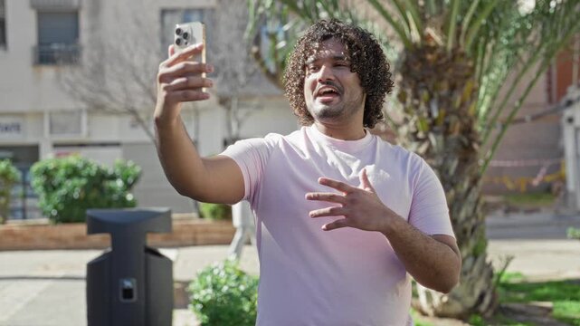 Man holding smartphone with raised arm and open palm while taking a selfie on a city street beside a palm tree and trash bin; confident.