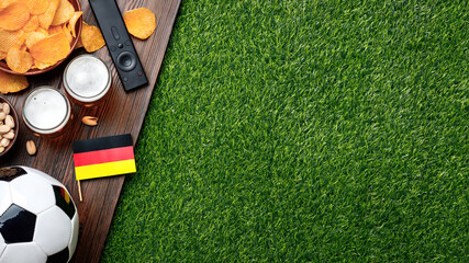 Naklejka premium Classic soccer ball, German mini flag, two glasses of beer, potato chips, pistachios and TV remote on wooden surface and green grass create the perfect game watching atmosphere. Copy space.
