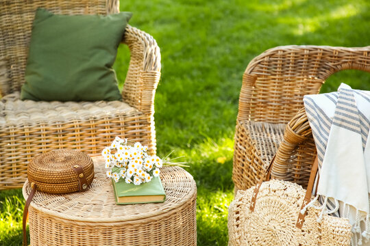 Comfortable wicker armchairs with chamomile flowers, book and bags on coffee table in park, outdoors