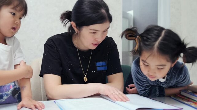 Asian mother guiding daughter reading textbook while second child waits nearby, focused home learning scene, parental support, education process, attention, literacy development, study routine
