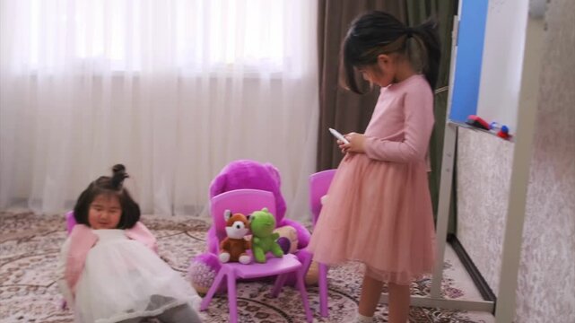 Kyrgyz Asian girls playing school at home, one child acting as teacher holding notebook while another lying and playing, contrasting behavior, discipline and learning concept, childhood interaction