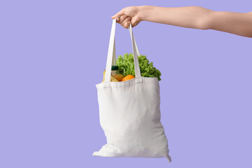 Female hand holding eco bag with different groceries on lilac background