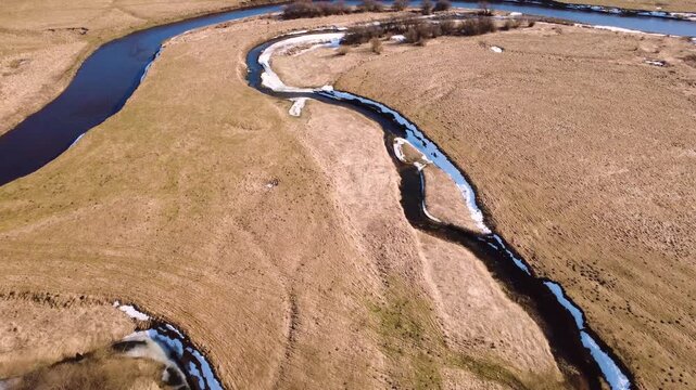 High-angle tracking forward shot over a dormant brown meadow featuring a meandering river, a small stream, and patches of melting snow in bright sunny daylight. High quality 4k aerial footage.