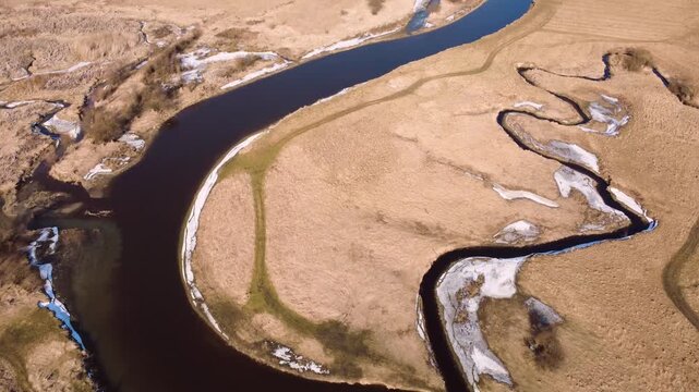 Aerial drone flight tracking over a winding dark river snaking through a dry, golden, late winter or early spring wilderness landscape with patches of melting snow. A beautiful cinematic view of untam