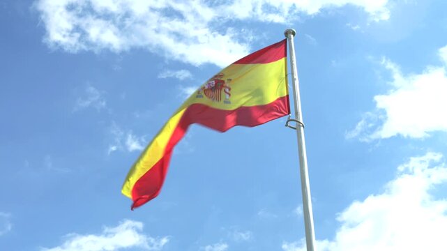 Spanish flag waving on tall pole against bright blue sky and clouds, patriotic national symbol of Spain in sunny weather