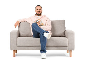 Fototapeta premium Young man with magazine sitting on grey sofa against white background
