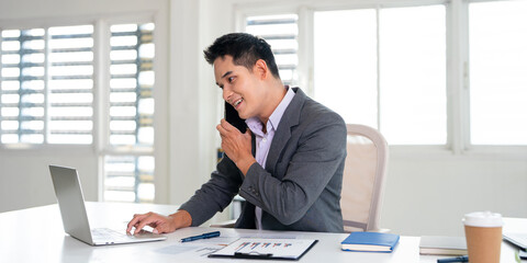 Fototapeta premium Businessman having call on smartphone with business partners or clients. Smiling mature man sitting at table talking by mobile cellphone at workplace in office.
