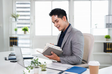 Fototapeta premium Businessman hand holding pen writing on notebook, work on laptop computer in modern office.