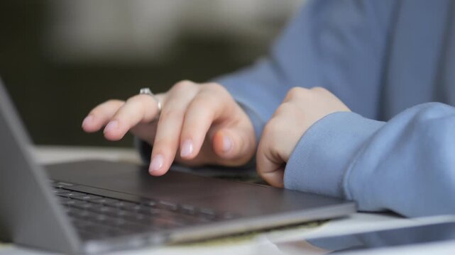 Close-up of hands typing on laptop keyboard. Fingers press keys with precision. Person wears blue long sleeve shirt. Ring visible on left hand. Background softly blurred