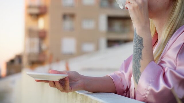 Woman in robe drinking coffee on terrace of modern apartment during sunrise, travel lifestyle concept, temporary stay, peaceful city exploration mood.