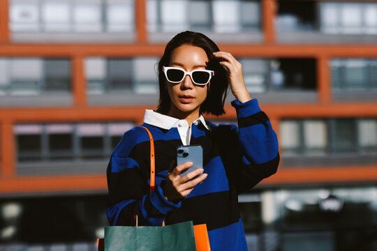 Asian woman wearing sunglasses and holding shopping bags using a smartphone outdoors