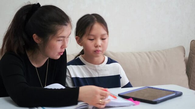 Kyrgyz Asian mother supervises young daughter doing homework with tablet and notebook, focused learning mixed with pressure, education stress and parental control in home environment