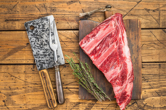 Fresh uncooked beef ribs slab with visible bones and herbs on cutting board. Wooden background for BBQ cooking and butchery concepts.