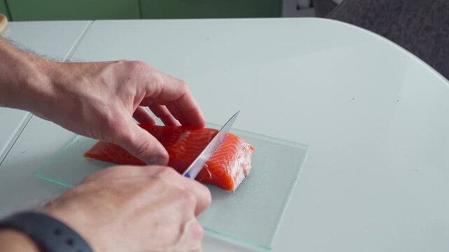Male hands slicing fresh salmon fillet on kitchen board with knife. Close-up of food preparation process, raw fish cutting for meal, healthy cooking, protein ingredient and home kitchen routine
