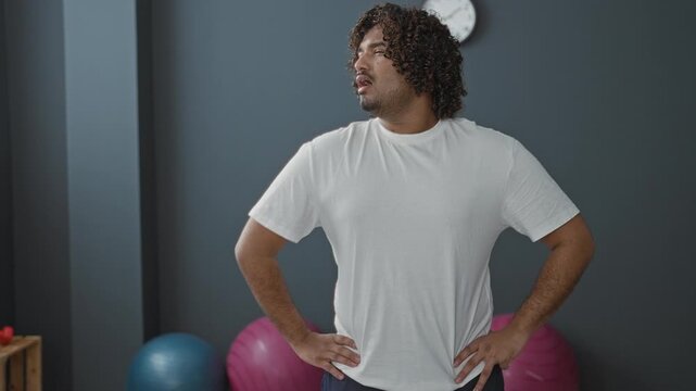 Young man wiping brow with hand and hands on hips beside exercise balls in a studio gym setting; post workout fatigue recovery.