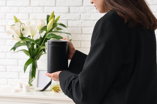 Young woman putting mortuary urn on chest of drawers near bouquet of funeral lily flowers in room, closeup