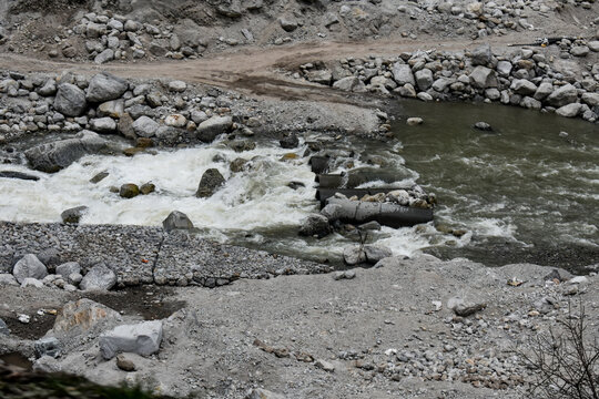 Fast-flowing mountain stream cutting through rocky terrain with scattered boulders and rugged dirt tracks, showcasing raw Himalayan landscape and natural erosion.