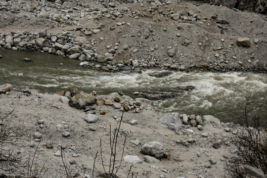 Fast-flowing mountain stream cutting through rocky terrain with scattered boulders and rugged dirt tracks, showcasing raw Himalayan landscape and natural erosion.