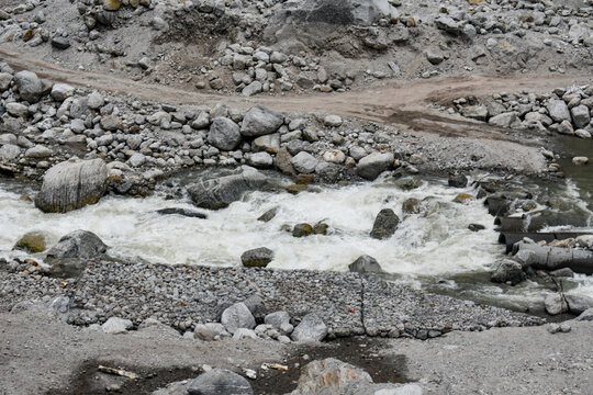 Fast-flowing mountain stream cutting through rocky terrain with scattered boulders and rugged dirt tracks, showcasing raw Himalayan landscape and natural erosion.