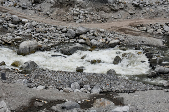 Fast-flowing mountain stream cutting through rocky terrain with scattered boulders and rugged dirt tracks, showcasing raw Himalayan landscape and natural erosion.