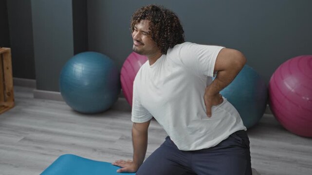 Man holding lower back on blue mat beside large exercise balls near wooden crate in a studio; discomfort recovery.