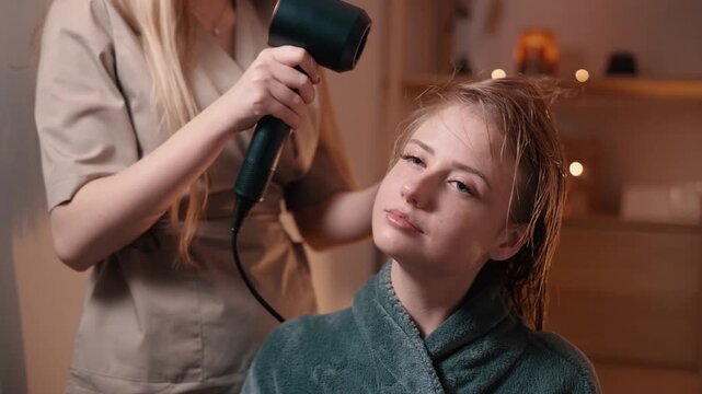 Professional hairstylist using hair dryer to style wet hair of young woman in cozy beauty salon, providing hair care and grooming service