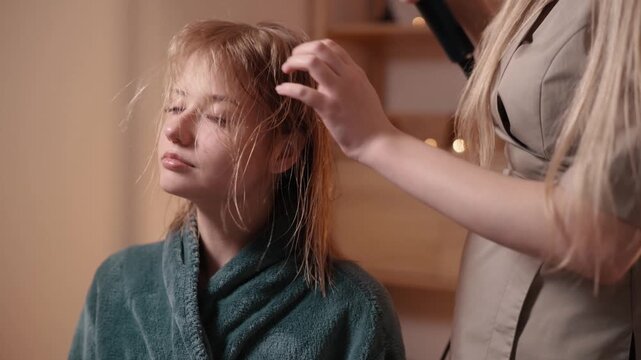 Professional hairstylist using hair dryer to style wet hair of young woman in cozy beauty salon, providing hair care and grooming service
