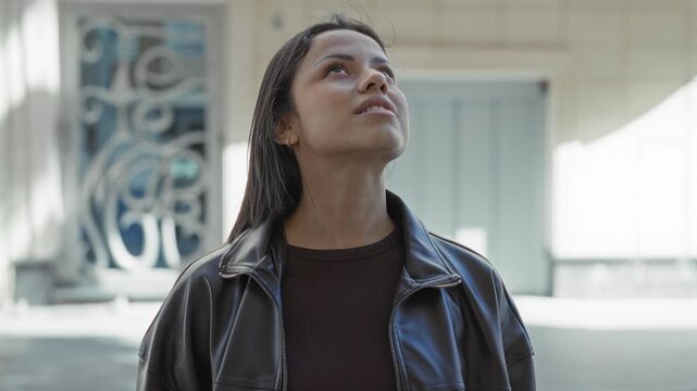Woman looking up showing bare neck and face, wearing leather jacket and brown sweater, standing by ornate building on street; contemplation.