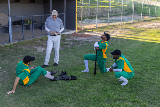 Coach holding two baseballs initiating batting practice while players adjusting gear near dugout