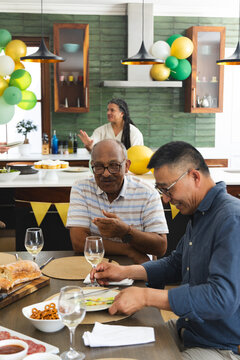 Diverse senior friends sharing meal in home kitchen at table with bread, cupcakes, balloons