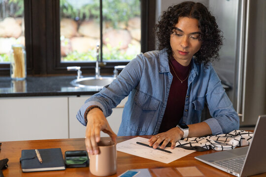 Non-binary adult reaching for ceramic mug while reviewing sketches with open laptop at wood counter