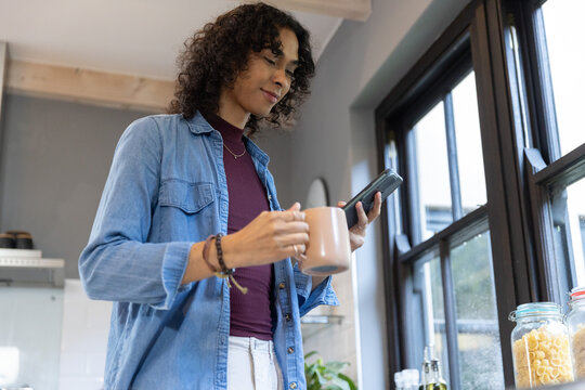Non-binary adult standing in kitchen wearing denim shirt, holding mug and checking phone near jars