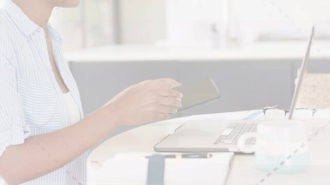 Woman at counter reaching for smartphone, tapping while shifting from laptop for business task