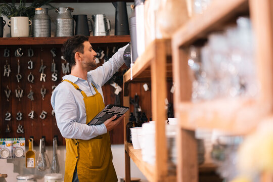 Shop worker stocking shelf in retail store interior, male staff arranging product on wooden shelf with apron