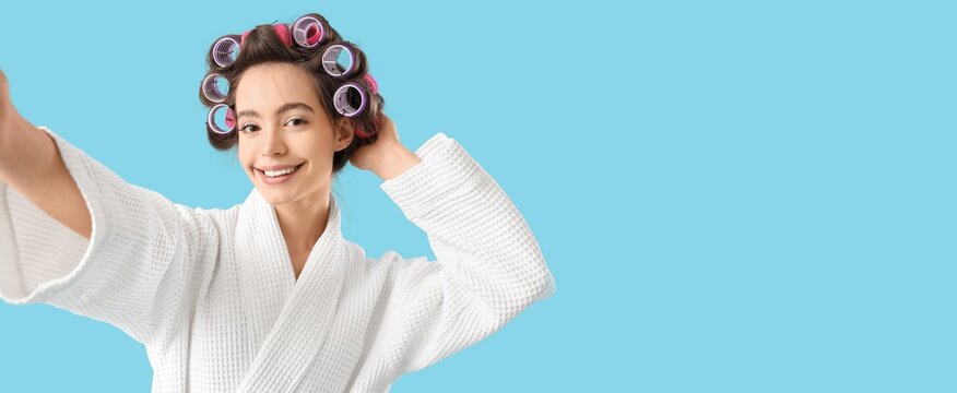 Young woman in bathrobe and with hair curlers taking selfie on light blue background with space for text